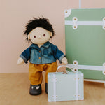 Olli ella dinkum doll stood on a wooden floor next to its sage travel tog case wearing a denim jacket and orange trousers
