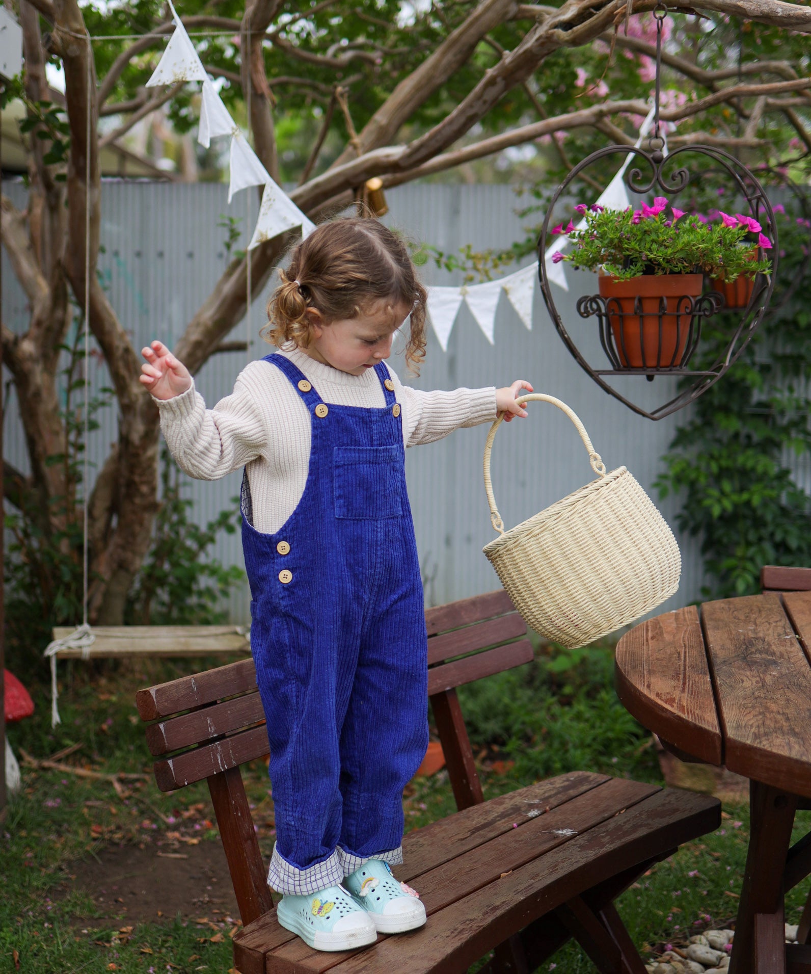 A child holding the Straw Olli Ella Rattan Gathering basket. The child is standing on a wooden bench. 