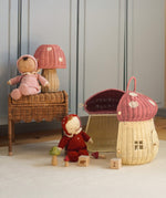 The Olli Ella Rattan Woven mushroom doll's house, with a pink spotty toadstool top and an opening door. Next to it are two toadstool dozy dinkum dolls and some toys. 