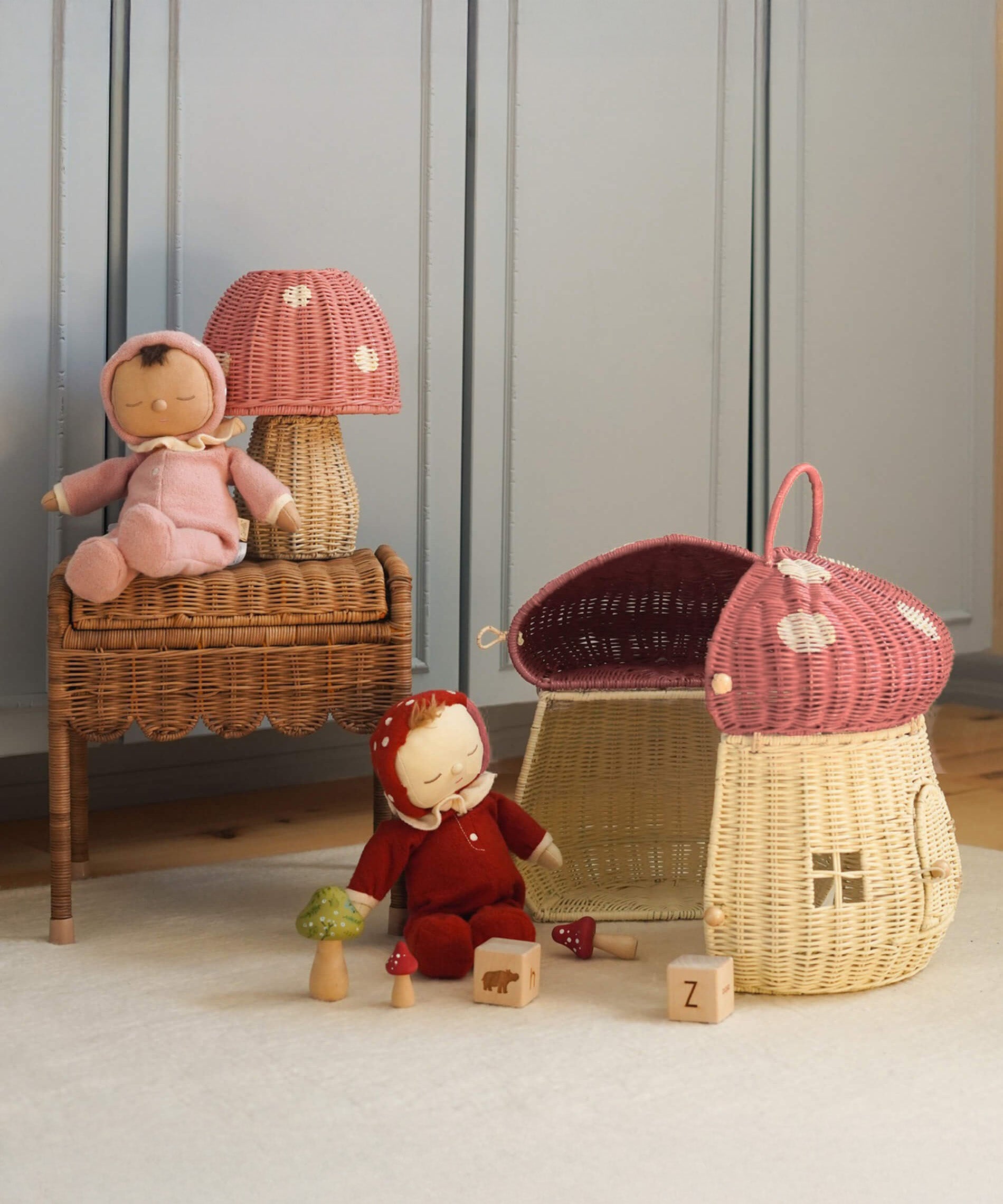 The Olli Ella Rattan Woven mushroom doll's house, with a pink spotty toadstool top and an opening door. Next to it are two toadstool dozy dinkum dolls and some toys. 