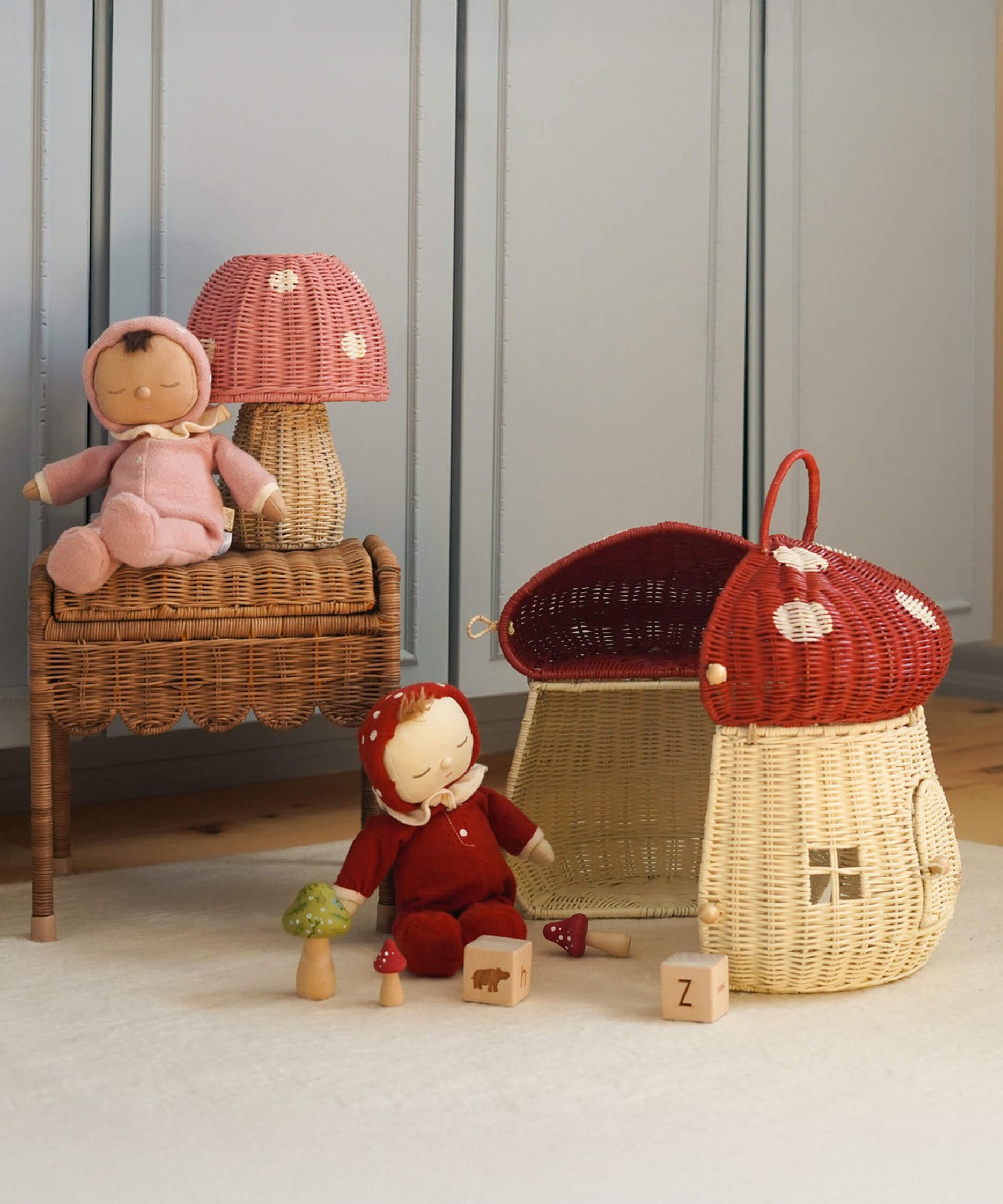 The Olli Ella Rattan Woven mushroom doll's house, with a red spotty toadstool top and an opening door. Next to it are two toadstool dozy dinkum dolls and some toys. 