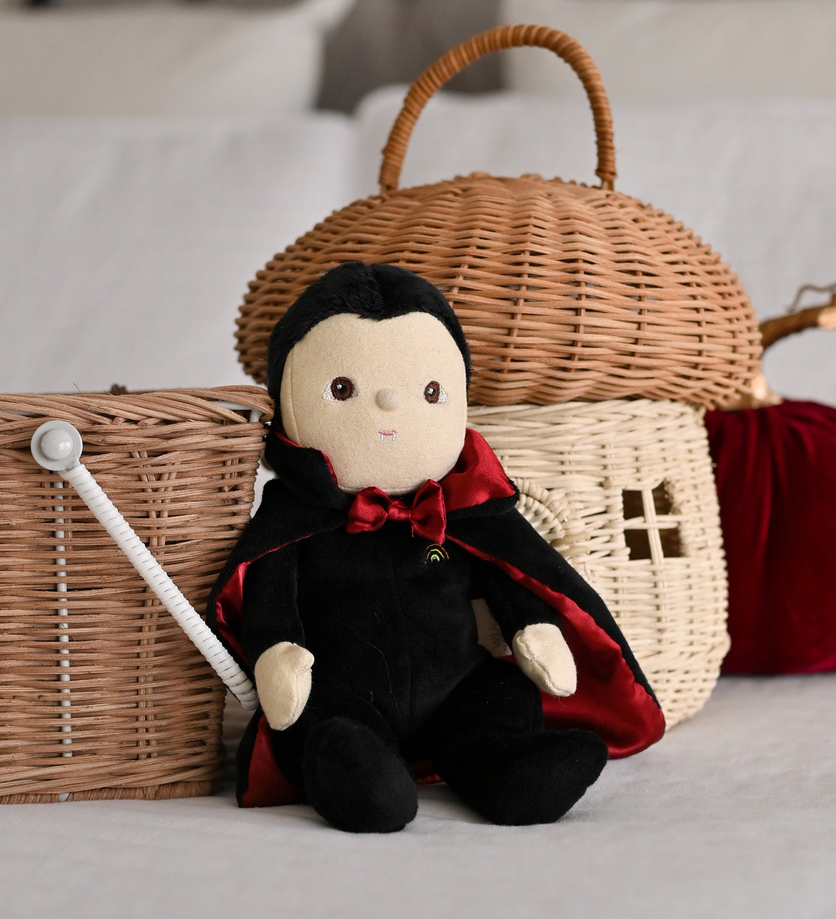 An Olli Ella Vincent Vampire Dinky Dinkum doll placed in a seated position in front of baskets. A soft vampire doll with a black cape with bat shaped glow in the dark details on the back. These soft toys are part of Olli Ella's Halloween collection available here at Babipur.