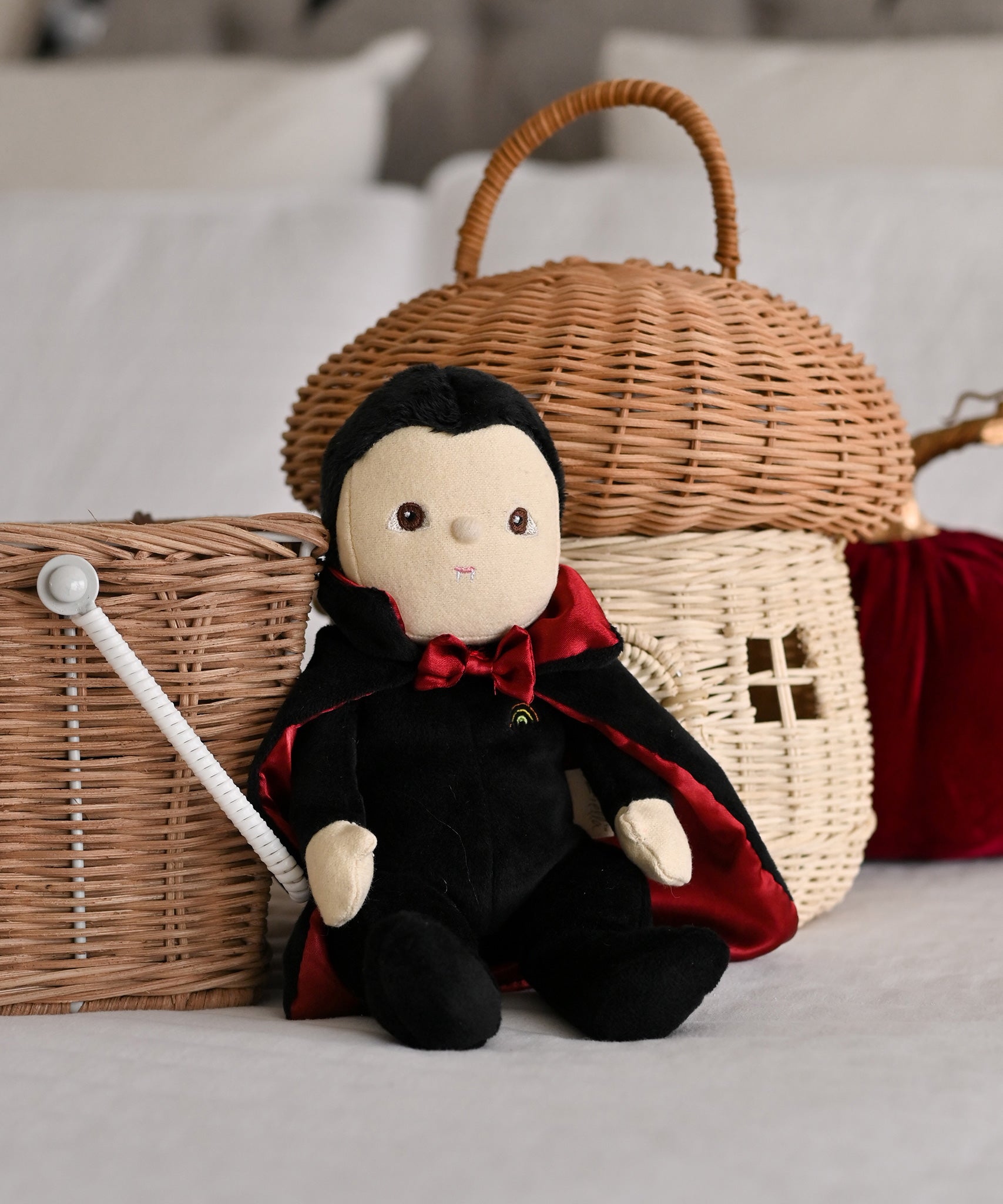 An Olli Ella Vincent Vampire Dinky Dinkum doll placed in a seated position in front of baskets. A soft vampire doll with a black cape with bat shaped glow in the dark details on the back. These soft toys are part of Olli Ella's Halloween collection available here at Babipur.