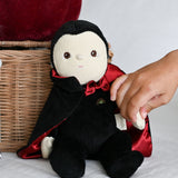 A child's hand reaching for the Olli Ella Vincent Vampire Dinky Dinkum doll. A soft vampire doll with a black cape with bat shaped glow in the dark details on the back. These soft toys are part of Olli Ella's Halloween collection available here at Babipur.