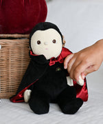 A child's hand reaching for the Olli Ella Vincent Vampire Dinky Dinkum doll. A soft vampire doll with a black cape with bat shaped glow in the dark details on the back. These soft toys are part of Olli Ella's Halloween collection available here at Babipur.