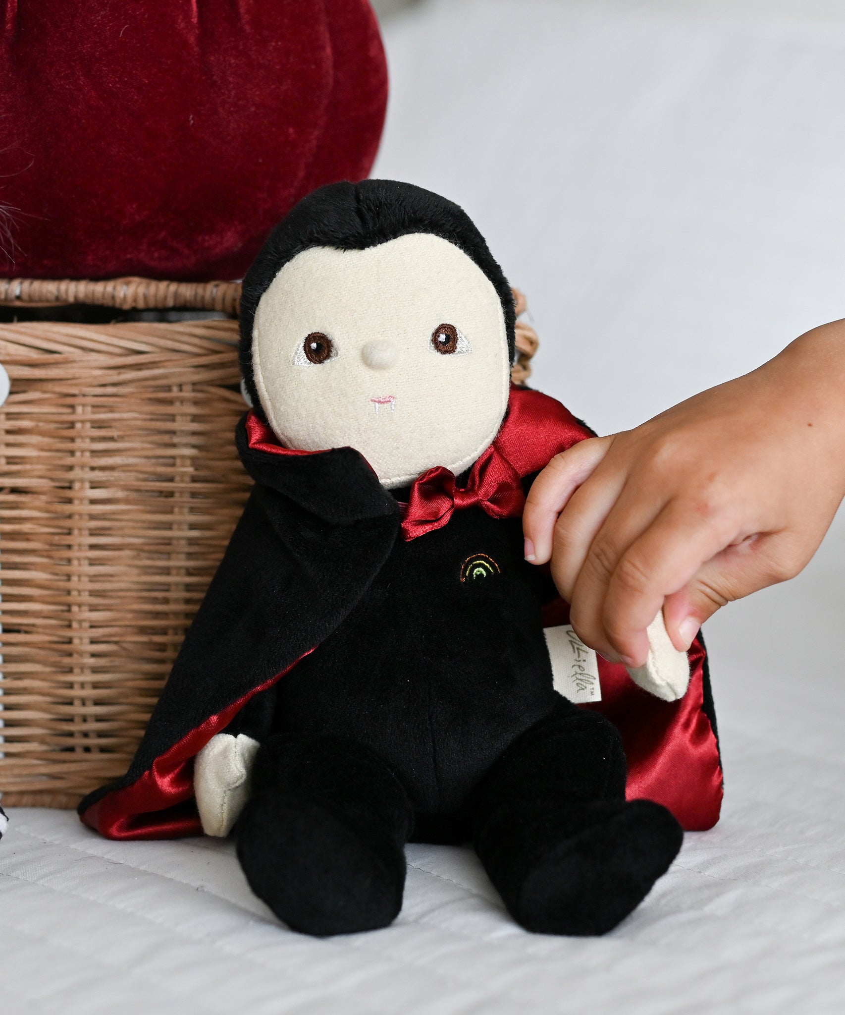 A child's hand reaching for the Olli Ella Vincent Vampire Dinky Dinkum doll. A soft vampire doll with a black cape with bat shaped glow in the dark details on the back. These soft toys are part of Olli Ella's Halloween collection available here at Babipur.