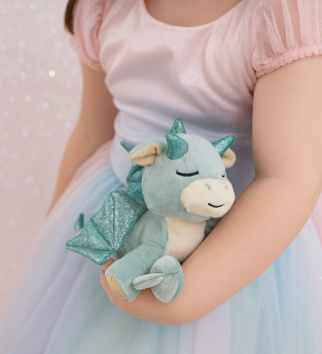 A child wearing a princess outfit and holding a Olli Ella misty volt dragon doll. This light green fluffy dragon has a sleeping face and features glittery wings and comes with a green magnetic pacifier. This soft toy is available at Babipur.