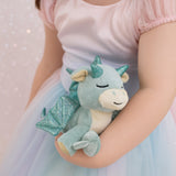 A child wearing a princess outfit and holding a Olli Ella misty volt dragon doll. This light green fluffy dragon has a sleeping face and features glittery wings and comes with a green magnetic pacifier. This soft toy is available at Babipur.