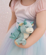 A child wearing a princess outfit and holding a Olli Ella misty volt dragon doll. This light green fluffy dragon has a sleeping face and features glittery wings and comes with a green magnetic pacifier. This soft toy is available at Babipur.