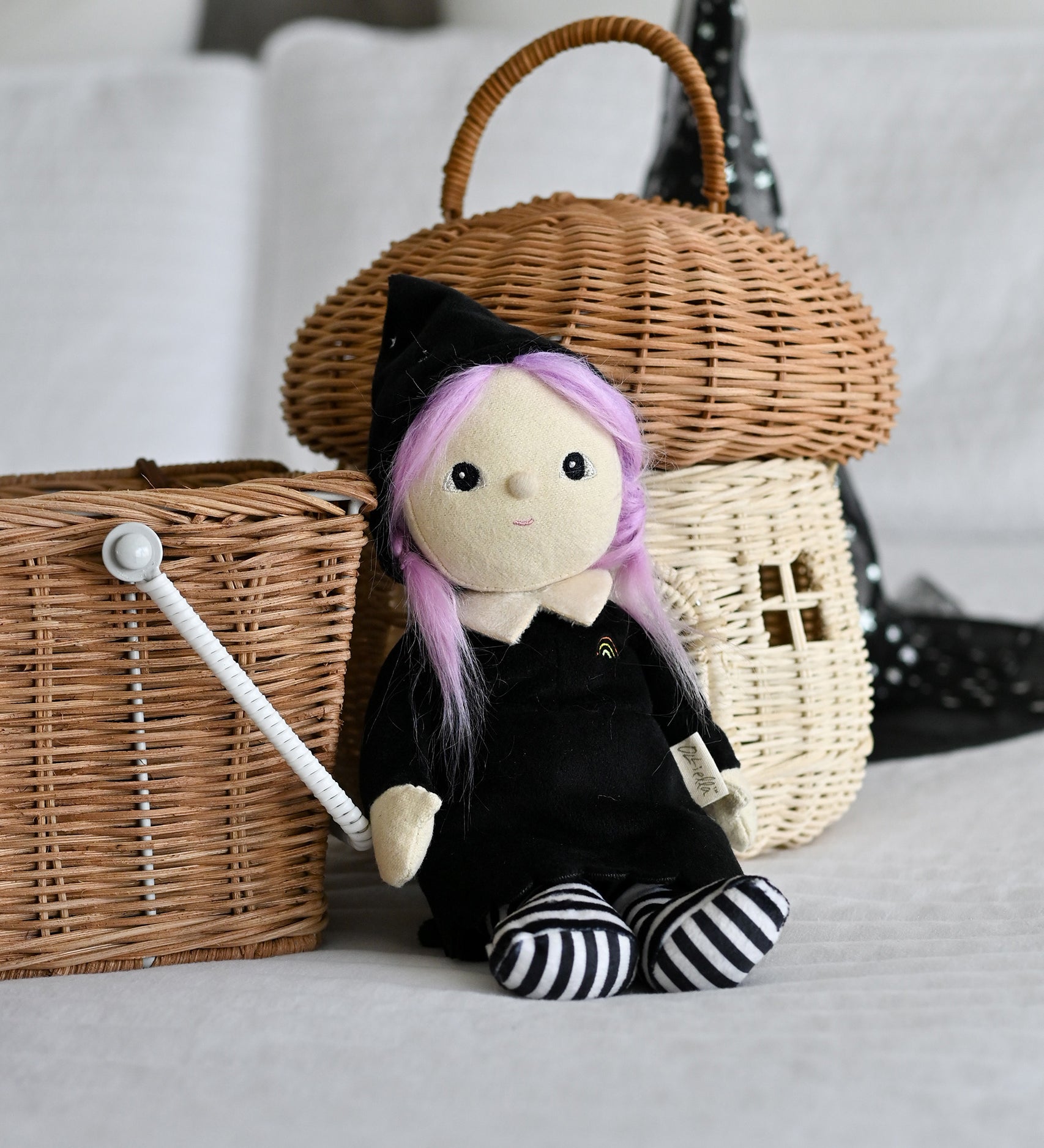 An Olli Ella Willa Witch Dinky Dinkum Doll placed in a seated position in front of baskets. A witch with a black outfit and matching hat with glow in the dark features on the striped tights, collar and stars on the hat. These soft toys are part of Olli Ella's Halloween collection available here at Babipur.