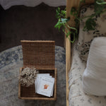 Olli Ella Petal Storie Stool in Natural Rattan. The stool has been placed by the side of a bed and has books placed inside, the lid has been left open.