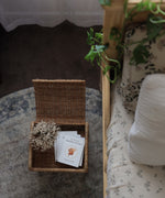 Olli Ella Petal Storie Stool in Natural Rattan. The stool has been placed by the side of a bed and has books placed inside, the lid has been left open.