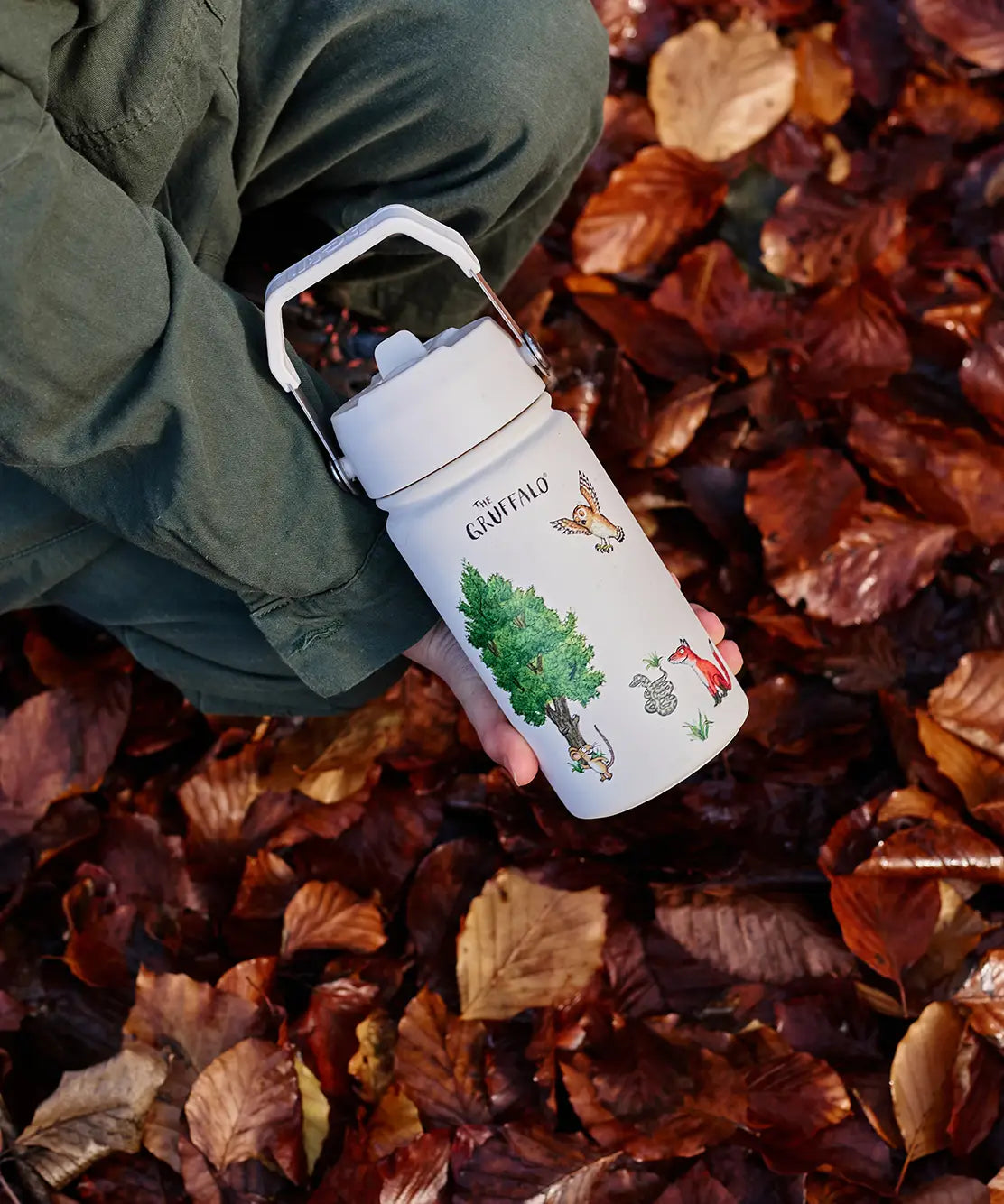 Child holding a One Green Bottle stainless steel water bottle in cream with Gruffalo characters above autumn leaves