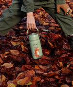 Child standing on Autumn leaves and holding a One Green Bottle stainless steel bottle in green with a Zog character