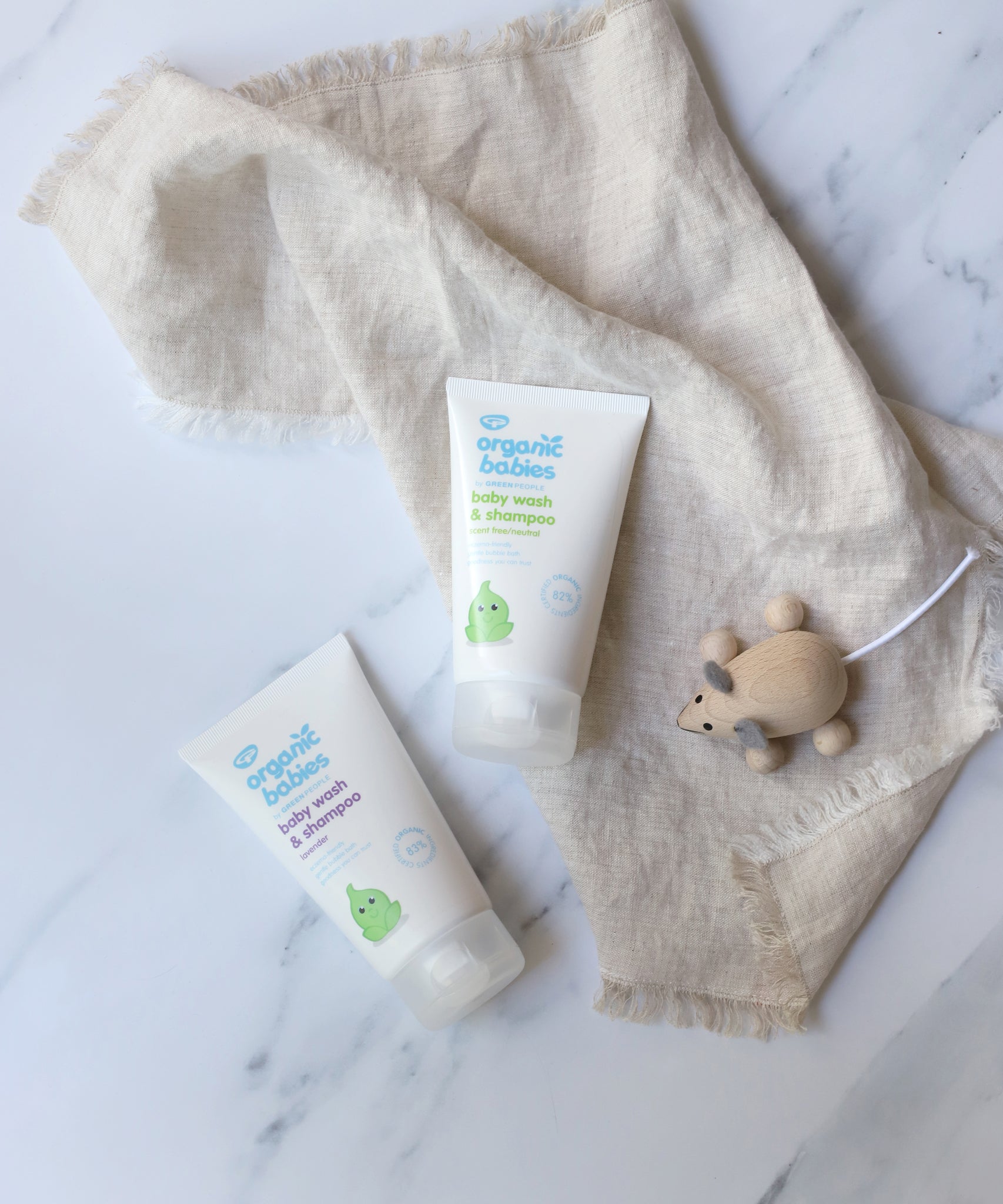 Green People Organic Babies unscented and lavender scented Baby Wash & Shampoo in 150ml tubes. The tubes have been placed on a linen coloured muslin cloth next to a wooden toy mouse. 