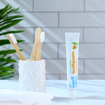 A tube of Organic Children Toothpaste Mandarin & Aloe Vera with Fluoride placed on a marble surface next to a ceramic pot containing bamboo toothbrushes. 