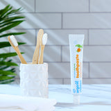 A tube of Organic Children Toothpaste Mandarin & Aloe Vera with Fluoride placed on a marble surface next to a ceramic pot containing bamboo toothbrushes. 