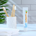 A tube of Organic Babies Toothpaste Mandarin & Aloe Vera pictured next to a pot of bamboo toothbrushes. A green leafy plant can be seen in the background. 