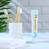 A tube of Organic Babies Toothpaste Mandarin & Aloe Vera pictured next to a pot of bamboo toothbrushes. A green leafy plant can be seen in the background. 