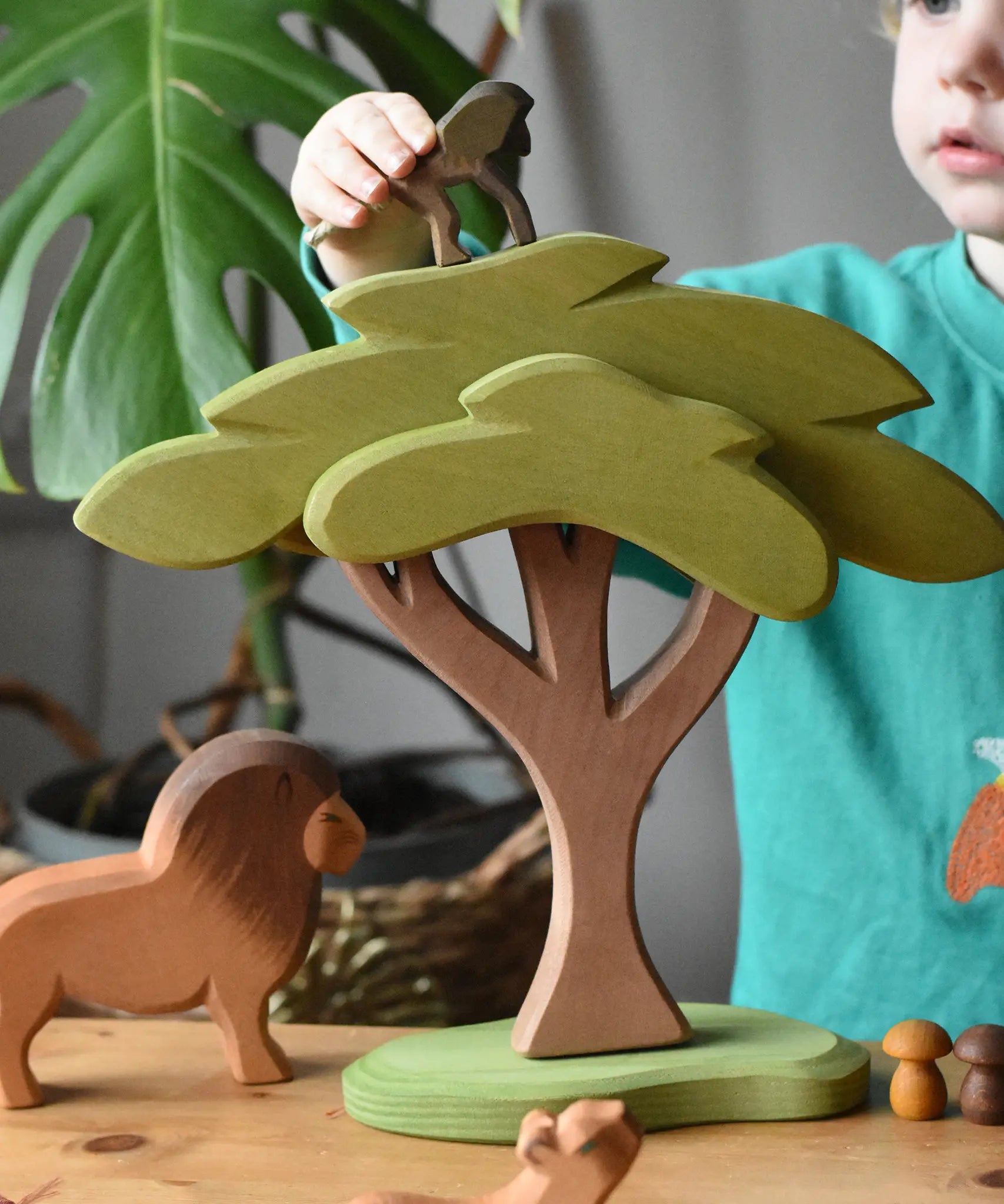 Ostheimer African tree wooden toy with support placed in a small world pay scene with lion figure A child is placing a standing baboon on top of the tree. 