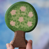 An Ostheimer wooden toy apple tree being held in an adult's hand to show scale. The apple tree has two decorated sides, this side shows the pink apple blossom flowers. This wooden figure is part of a large range of expertly hand crafted, Waldorf inspired, wooden toys here at Babipur. 