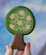 An Ostheimer wooden toy apple tree being held in an adult's hand to show scale. The apple tree has two decorated sides, this side shows the pink apple blossom flowers. This wooden figure is part of a large range of expertly hand crafted, Waldorf inspired, wooden toys here at Babipur. 