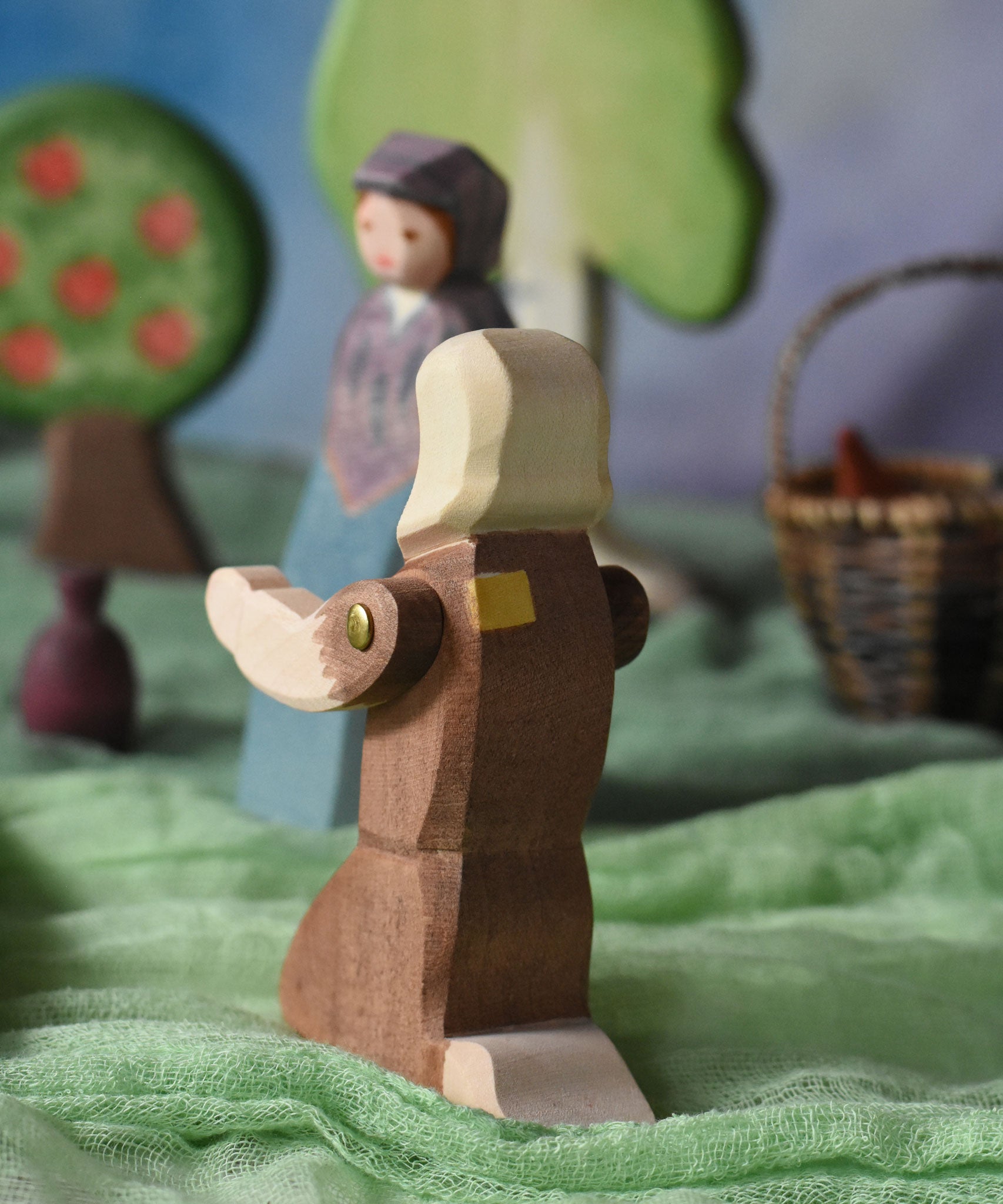 A view of the back of the Ostheimer beggar wooden toy figure placed in a small world play scene. The figure has moveable arms and is wearing a raggedy brown outfit with holes and patches on. This wooden figure is part of a large range of expertly hand crafted, Waldorf inspired, wooden toys here at Babipur. 