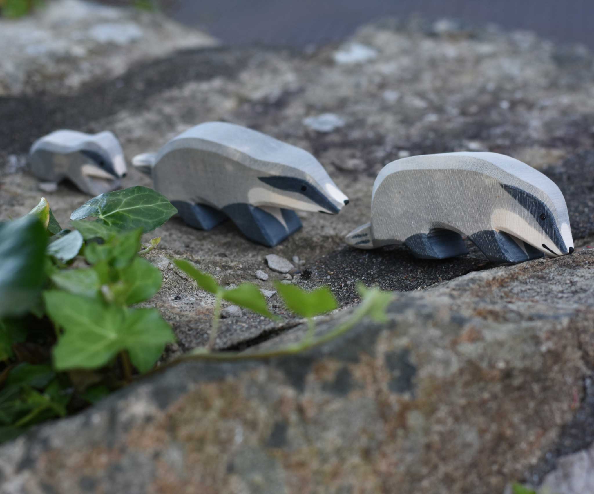 A group of three wooden toy badgers lined up on a stone wall outdoors. The one at the front has it's head down, the one in the middle has it's head straight with the small badger following at the back of the line.  