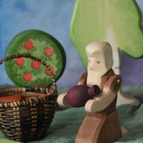 An Ostheimer beggar wooden toy figure placed in a small world play scene. The figure has moveable arms and is wearing a raggedy brown outfit with holes and patches on. The Beggar has his arms placed out and is holding a purple Grapat mandala piece in this arms. This wooden figure is part of a large range of expertly hand crafted, Waldorf inspired, wooden toys here at Babipur. 