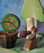 An Ostheimer beggar wooden toy figure placed in a small world play scene. The figure has moveable arms and is wearing a raggedy brown outfit with holes and patches on. The Beggar has his arms placed out and is holding a purple Grapat mandala piece in this arms. This wooden figure is part of a large range of expertly hand crafted, Waldorf inspired, wooden toys here at Babipur. 