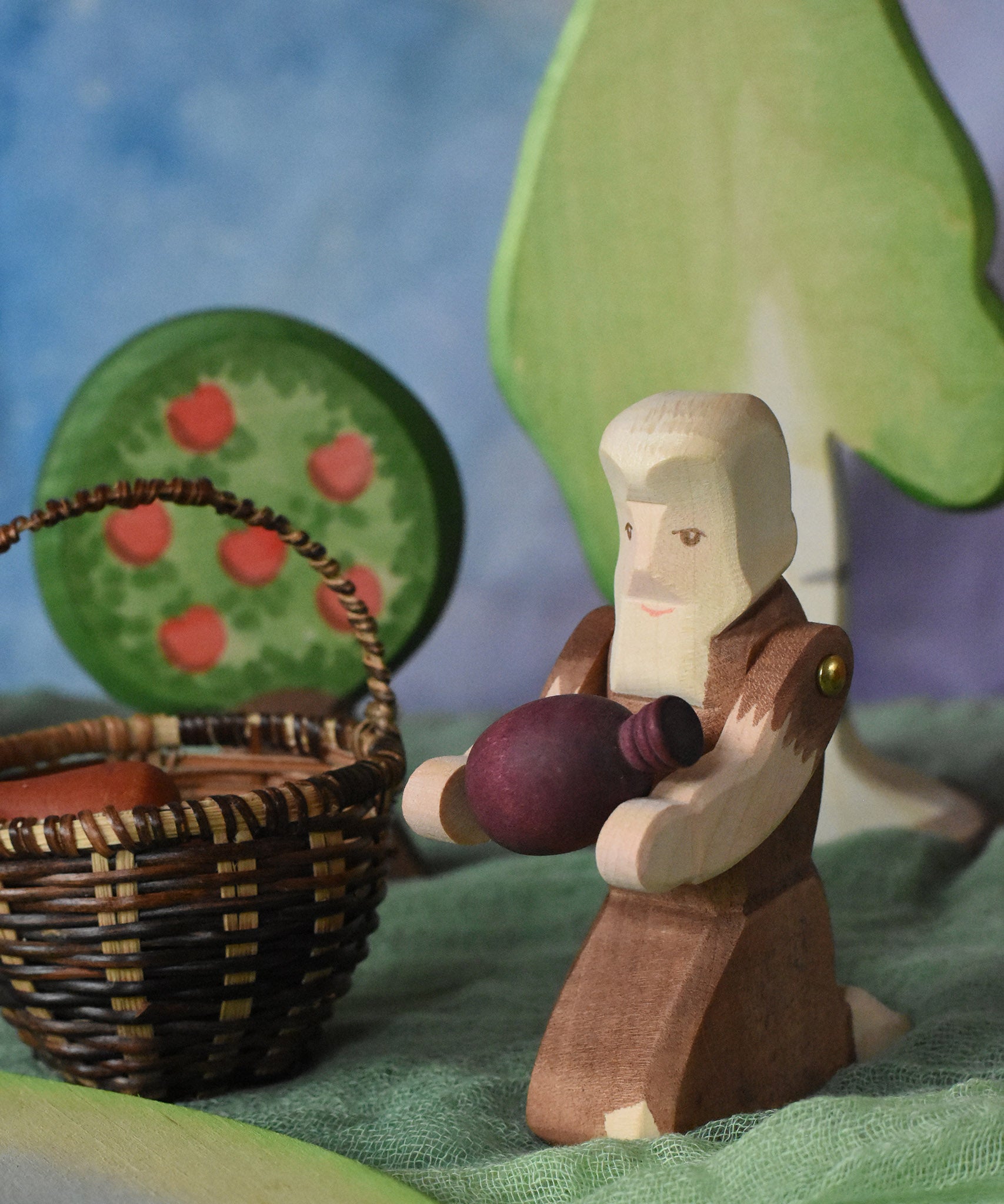 An Ostheimer beggar wooden toy figure placed in a small world play scene. The figure has moveable arms and is wearing a raggedy brown outfit with holes and patches on. The Beggar has his arms placed out and is holding a purple Grapat mandala piece in this arms. This wooden figure is part of a large range of expertly hand crafted, Waldorf inspired, wooden toys here at Babipur. 