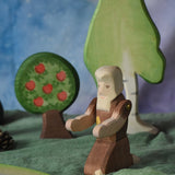 An Ostheimer beggar wooden toy figure placed in a small world play scene. The figure has moveable arms and is wearing a raggedy brown outfit with holes and patches on. There is an Ostheimer apple tree and a large birch tree in the background. This wooden figure is part of a large range of expertly hand crafted, Waldorf inspired, wooden toys here at Babipur. 