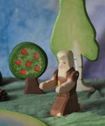 An Ostheimer beggar wooden toy figure placed in a small world play scene. The figure has moveable arms and is wearing a raggedy brown outfit with holes and patches on. There is an Ostheimer apple tree and a large birch tree in the background. This wooden figure is part of a large range of expertly hand crafted, Waldorf inspired, wooden toys here at Babipur. 