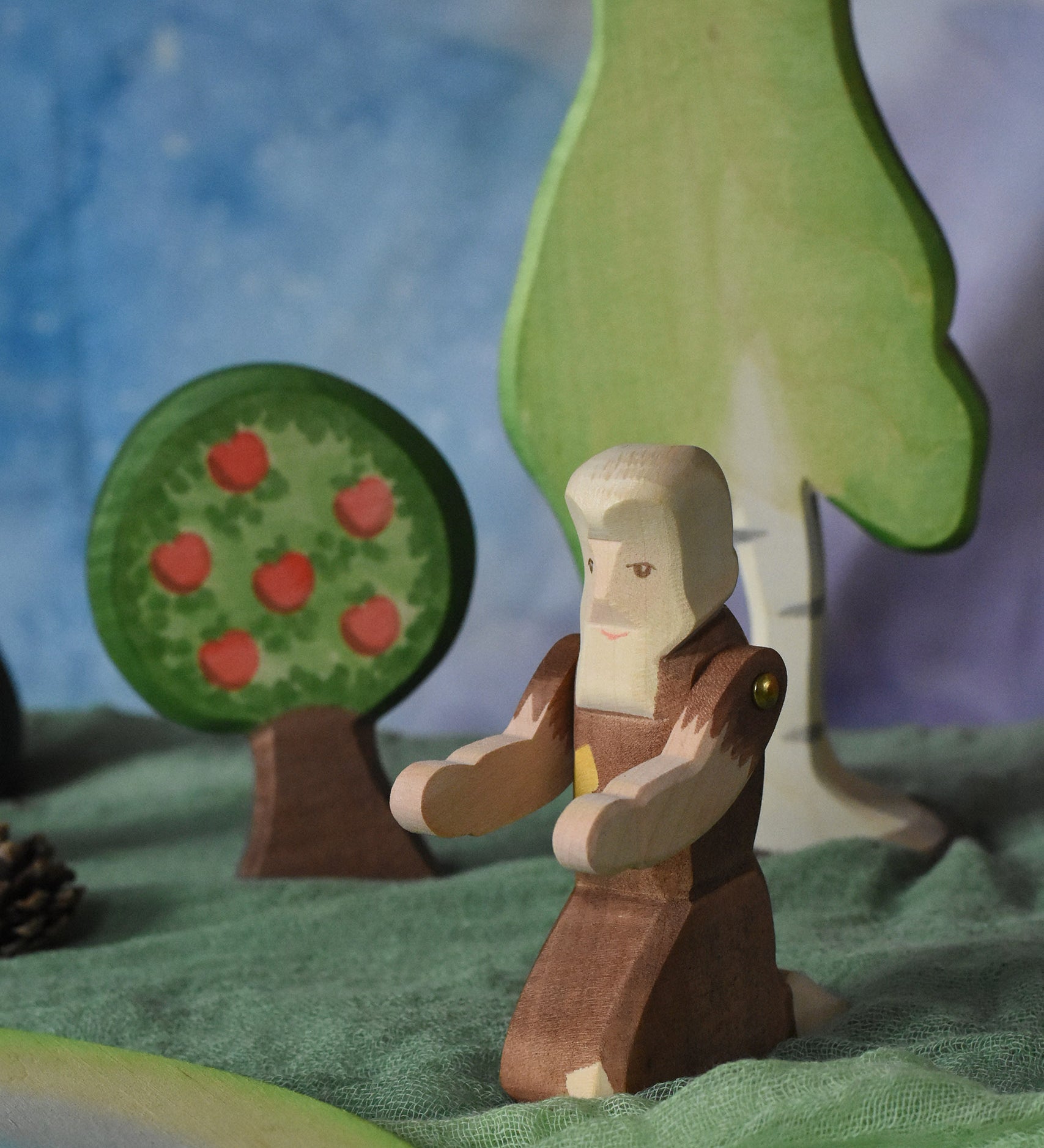 An Ostheimer beggar wooden toy figure placed in a small world play scene. The figure has moveable arms and is wearing a raggedy brown outfit with holes and patches on. There is an Ostheimer apple tree and a large birch tree in the background. This wooden figure is part of a large range of expertly hand crafted, Waldorf inspired, wooden toys here at Babipur. 