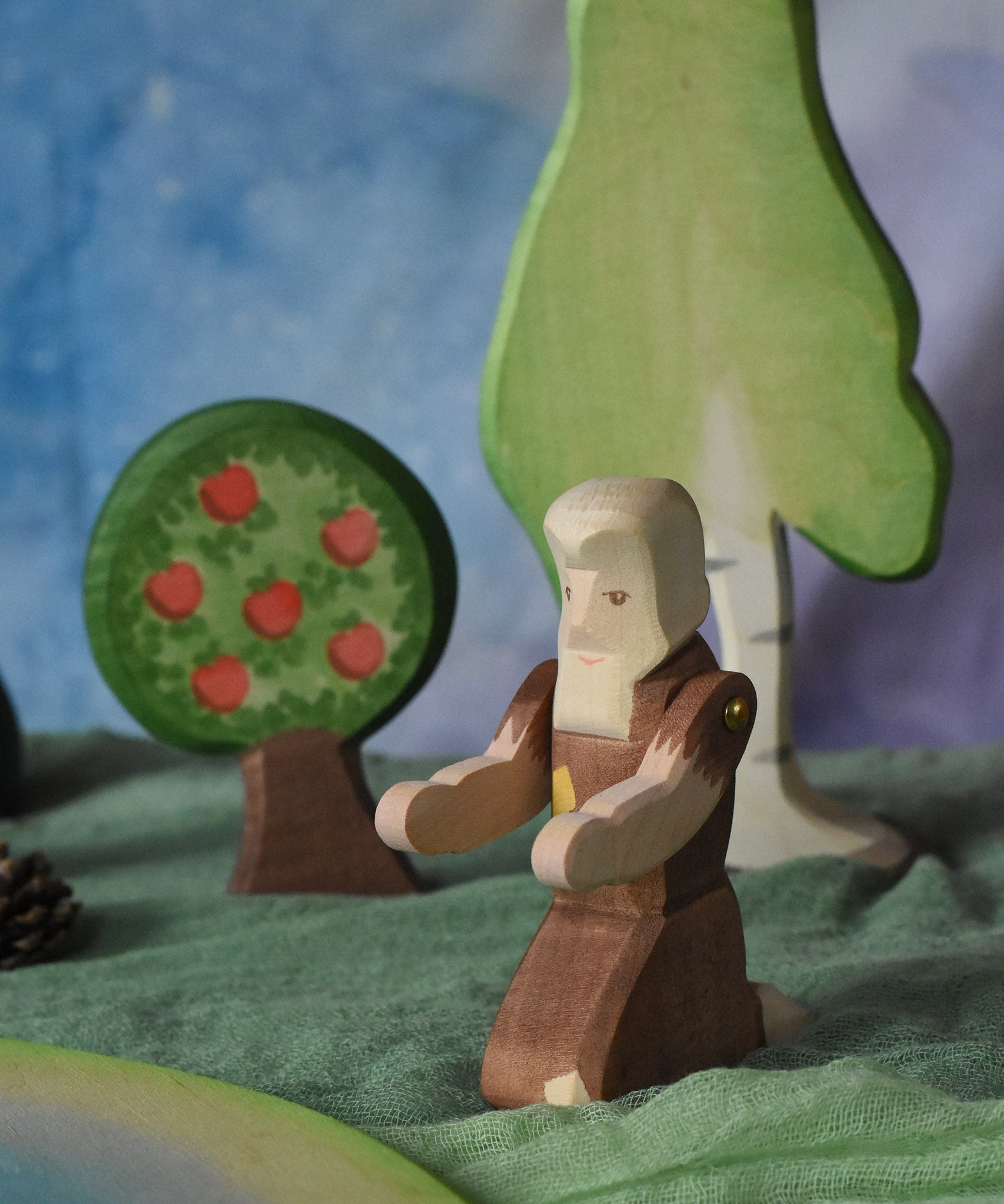 An Ostheimer beggar wooden toy figure placed in a small world play scene. The figure has moveable arms and is wearing a raggedy brown outfit with holes and patches on. There is an Ostheimer apple tree and a large birch tree in the background. This wooden figure is part of a large range of expertly hand crafted, Waldorf inspired, wooden toys here at Babipur. 