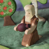 An Ostheimer beggar wooden toy figure placed in a small world play scene. The figure has moveable arms and is wearing a raggedy brown outfit with holes and patches on. The Beggar has his arms held out and is holding a purple Grapat mandala piece in this arms. This wooden figure is part of a large range of expertly hand crafted, Waldorf inspired, wooden toys here at Babipur. 