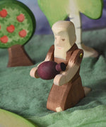 An Ostheimer beggar wooden toy figure placed in a small world play scene. The figure has moveable arms and is wearing a raggedy brown outfit with holes and patches on. The Beggar has his arms held out and is holding a purple Grapat mandala piece in this arms. This wooden figure is part of a large range of expertly hand crafted, Waldorf inspired, wooden toys here at Babipur. 