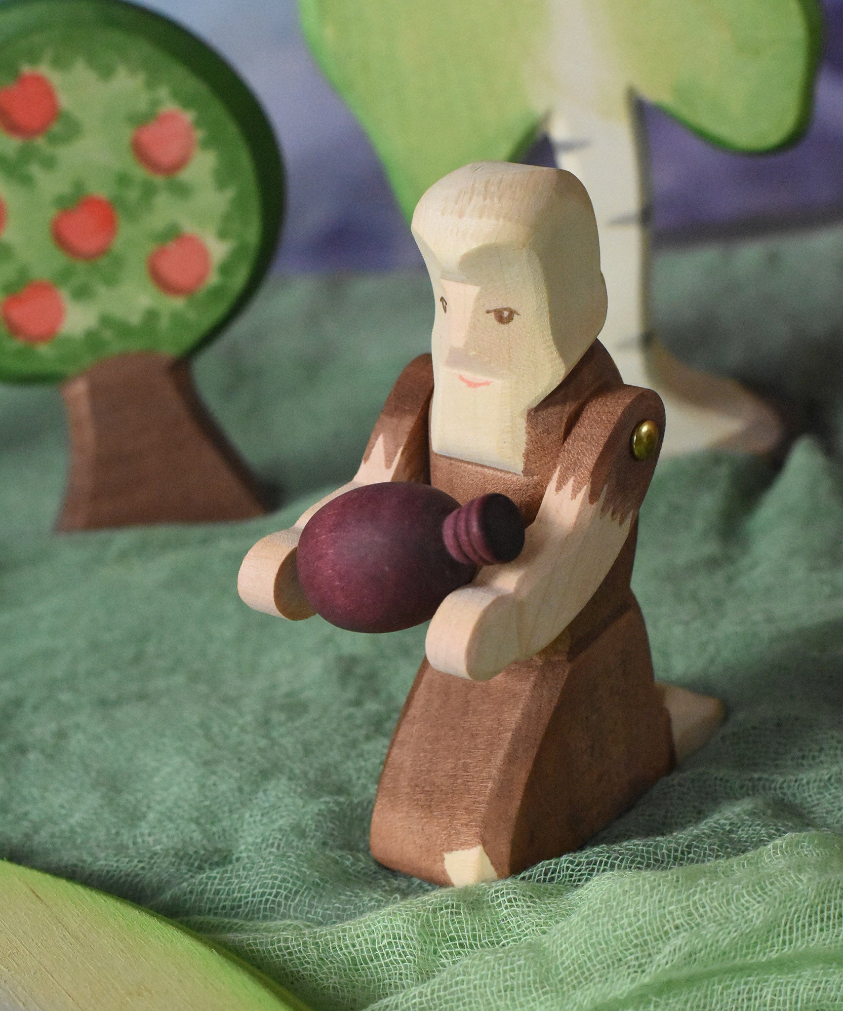 An Ostheimer beggar wooden toy figure placed in a small world play scene. The figure has moveable arms and is wearing a raggedy brown outfit with holes and patches on. The Beggar has his arms held out and is holding a purple Grapat mandala piece in this arms. This wooden figure is part of a large range of expertly hand crafted, Waldorf inspired, wooden toys here at Babipur. 