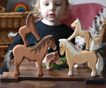 Two Ostheimer colts wooden toy horse figures placed on the back of two larger wooden toy horses. Child can be seen looking on on the background. 
