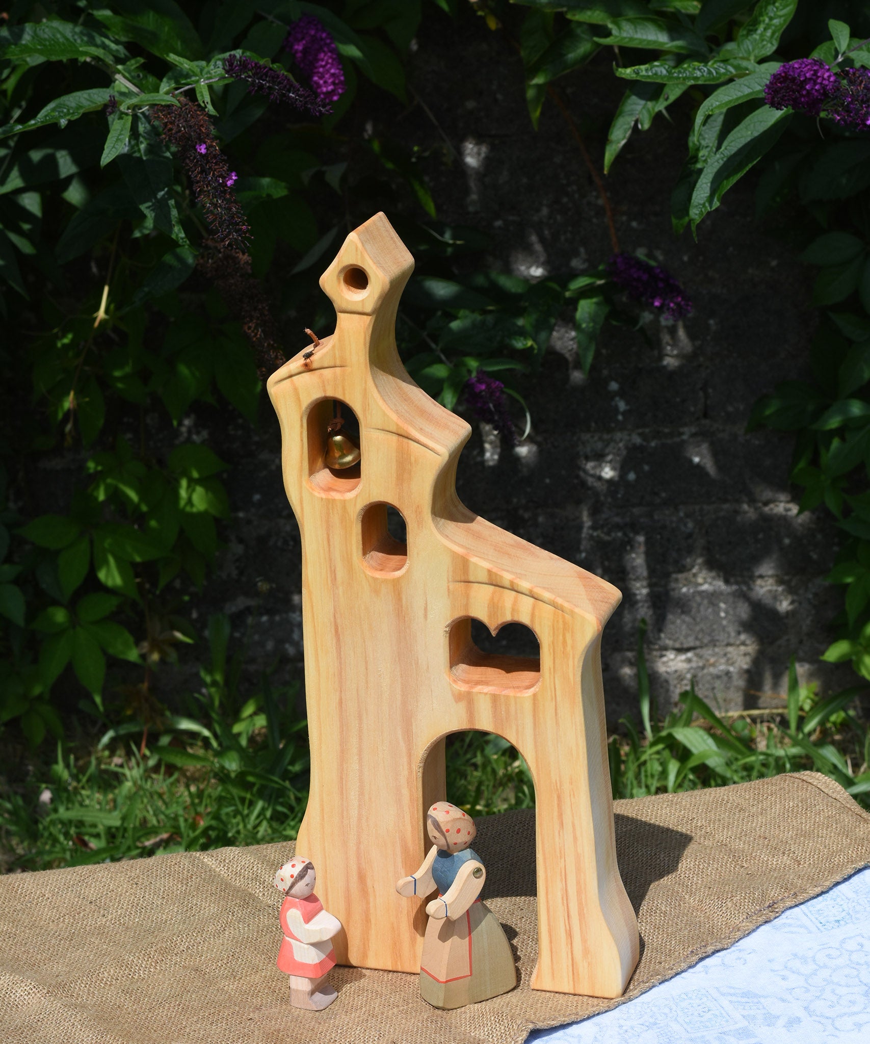 An Ostheimer wooden bell tower placed on a small table outdoors. These wooden figures are part of a large range of expertly hand crafted, Waldorf inspired, wooden toys here at Babipur.