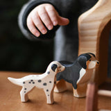 A child's hand reaching for Ostheimer dog figures that have been placed on a wooden surface. There is a dalmatian and a Bernese mountain dog.  These wooden figures are part of a wide range of expertly hand crafted wooden toys here at Babipur. 