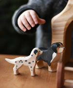 A child's hand reaching for Ostheimer dog figures that have been placed on a wooden surface. There is a dalmatian and a Bernese mountain dog.  These wooden figures are part of a wide range of expertly hand crafted wooden toys here at Babipur. 