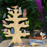 The Ostheimer wooden bird tree shown with various garden birds and owls placed on it's branches. The tree is placed on a jute covered surface besides a pile of books which have more Ostheimer birds placed on them.
