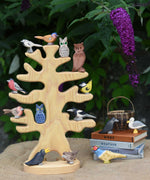 The Ostheimer wooden bird tree shown with various garden birds and owls placed on it's branches. The tree is placed on a jute covered surface besides a pile of books which have more Ostheimer birds placed on them.