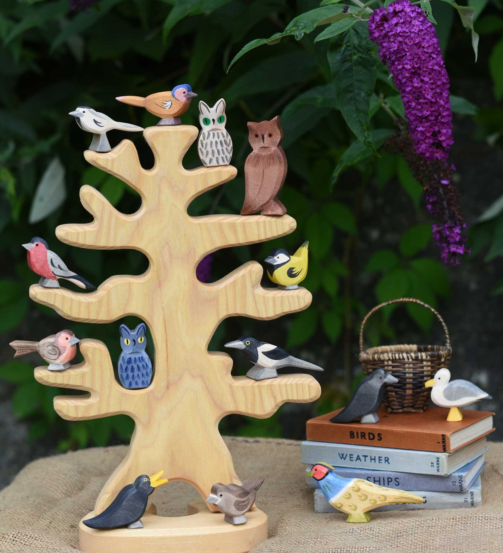 The Ostheimer wooden bird tree shown with various garden birds and owls placed on it's branches. The tree is placed on a jute covered surface besides a pile of books which have more Ostheimer birds placed on them.