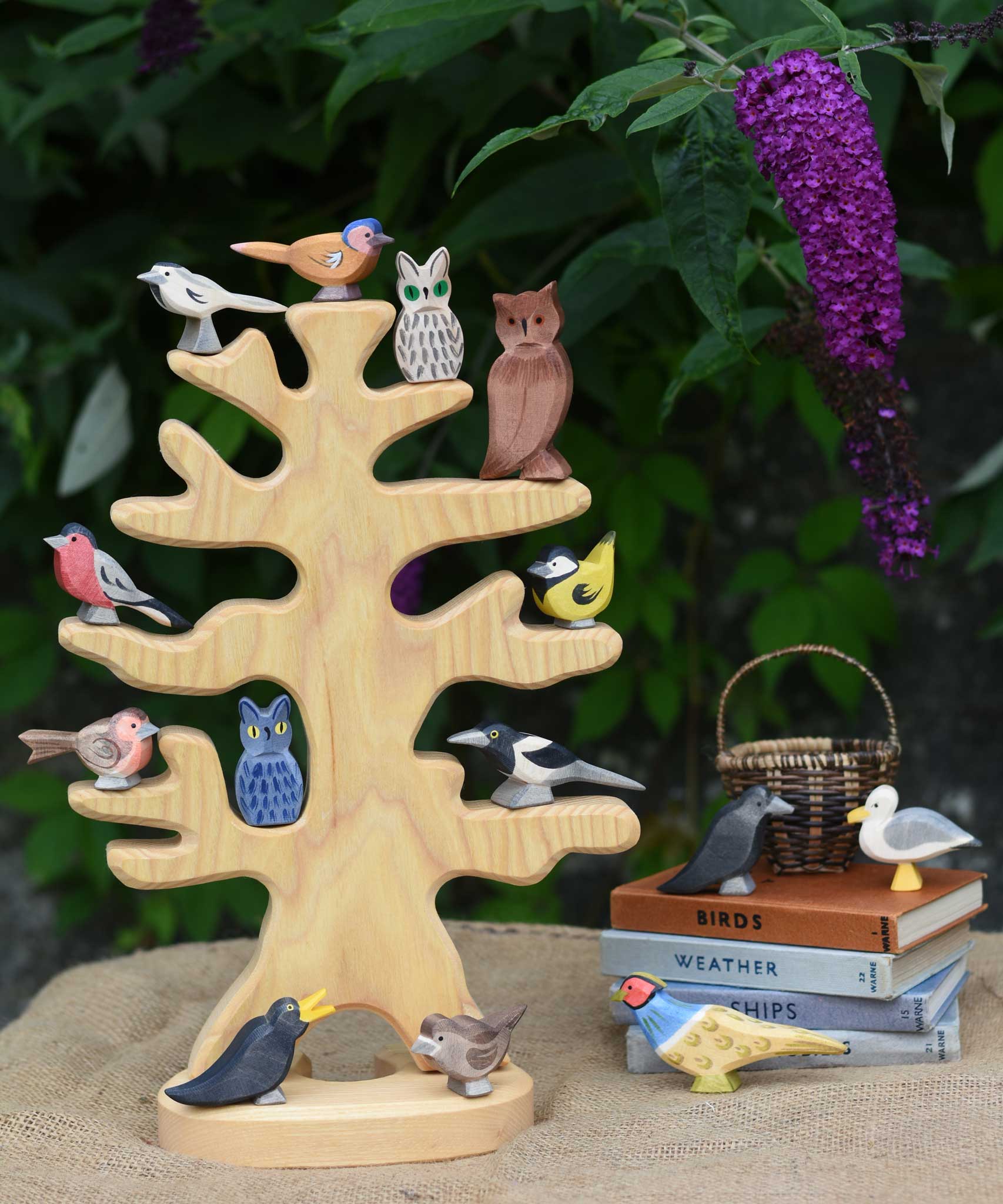 The Ostheimer wooden bird tree shown with various garden birds and owls placed on it's branches. The tree is placed on a jute covered surface besides a pile of books which have more Ostheimer birds placed on them.