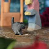 An Ostheimer hand made and hand painted wooden black cat figures  placed on a wooden surface. An Ostheimer wooden well and witch can be seen in the background. These wooden figures are part of a wide range of expertly hand crafted wooden toys here at Babipur. 