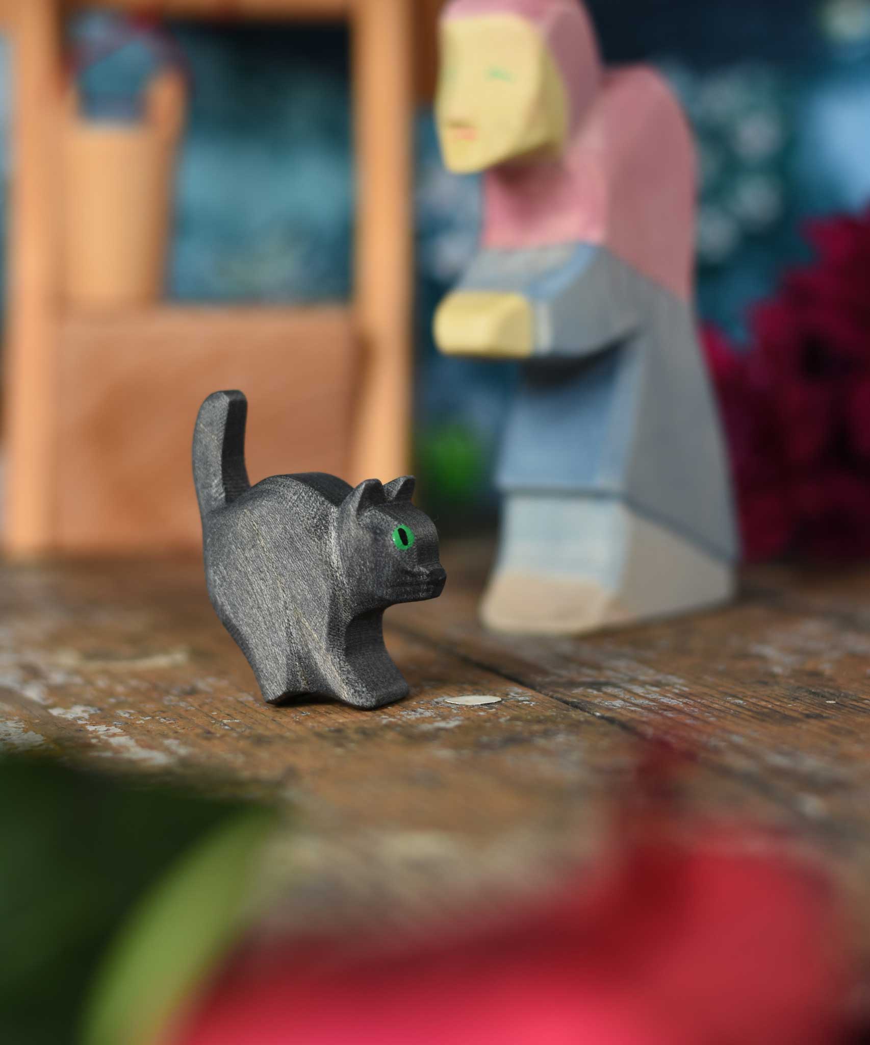 An Ostheimer hand made and hand painted wooden black cat figures  placed on a wooden surface. An Ostheimer wooden well and witch can be seen in the background. These wooden figures are part of a wide range of expertly hand crafted wooden toys here at Babipur. 