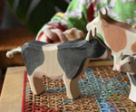 Ostheimer black and white cow wooden toy figure placed on a rattan stool in the middle of a simple small world play scene featuring cows and calves.