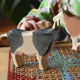 Ostheimer black and white cow wooden toy figure placed on a rattan stool in the middle of a simple small world play scene featuring cows and calves.